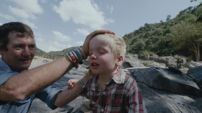 A father washes his sons face near a river surrounded by large boulders