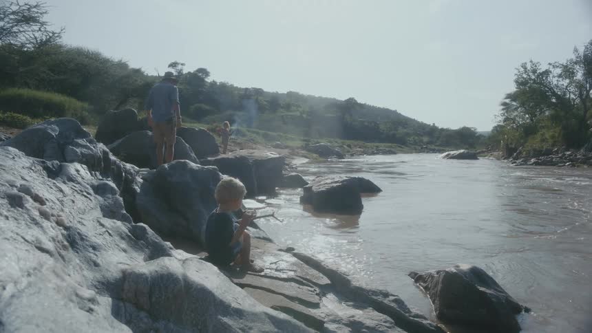 Father with hat blue shirt and shorts walks on river bank next to blonde son
