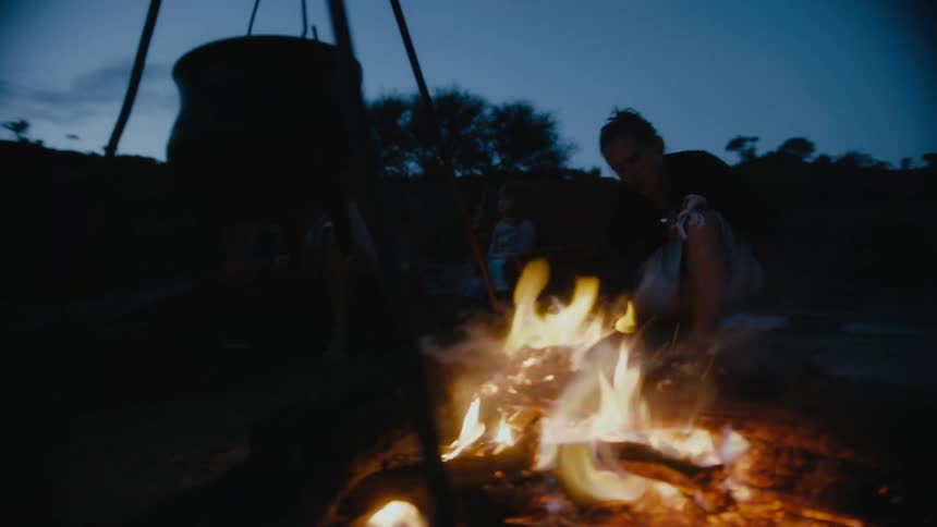 A woman boils water over a campfire near the woods