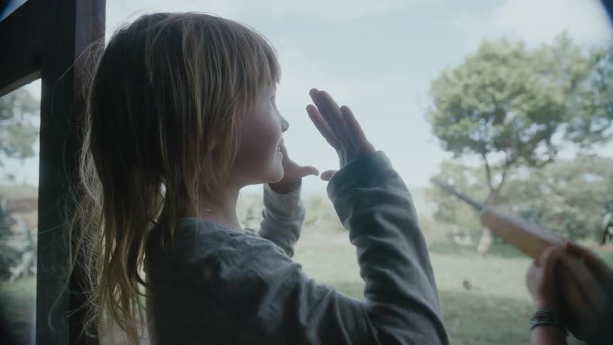 A young girl on farmland holds her hands in the air as her little brother points a toy gun off in to the distance and their mother watches them