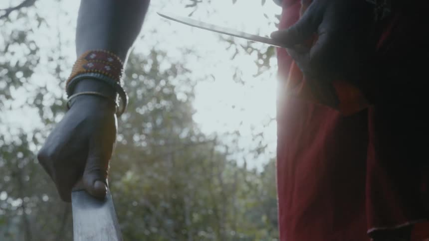 Black person sharpens a knife outdoors near the forest by sliding it over the blade of another knife