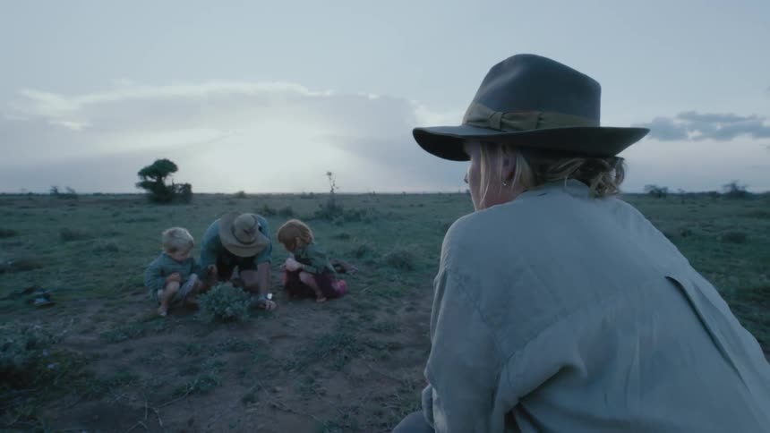 Boy and girl crouch on each side of their father as he digs in a field while the mother watches from a distance