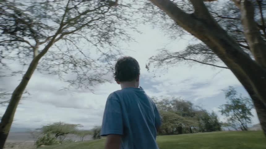 Young boy walks past a tree in a grassy field then turns and runs past the tree