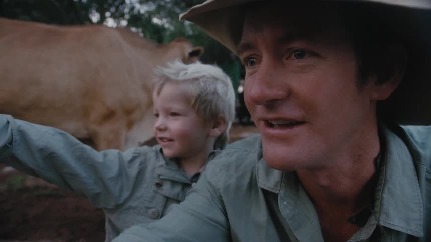 A man and his son crouch on the ground pointing while standing next to a cow