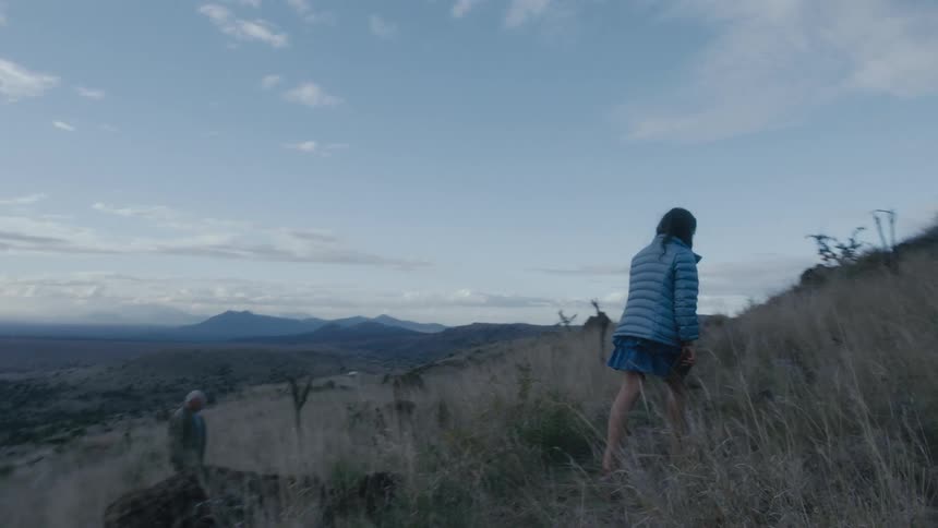 Grandfather and granddaughter walk along slope of mountain with mountains in distance
