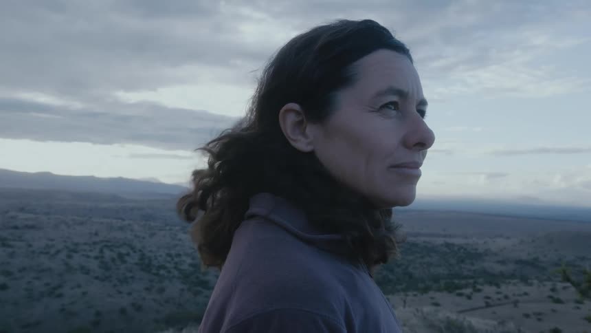 Dark haired woman wearing a jacket stands outdoors with a field and mountains behind her