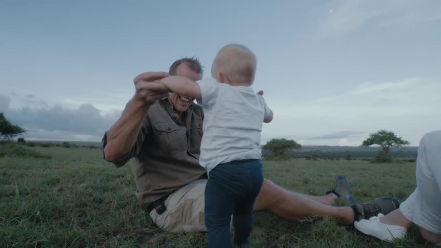 Grandfather sitting on a grass field helping his baby grandson stand up and walk