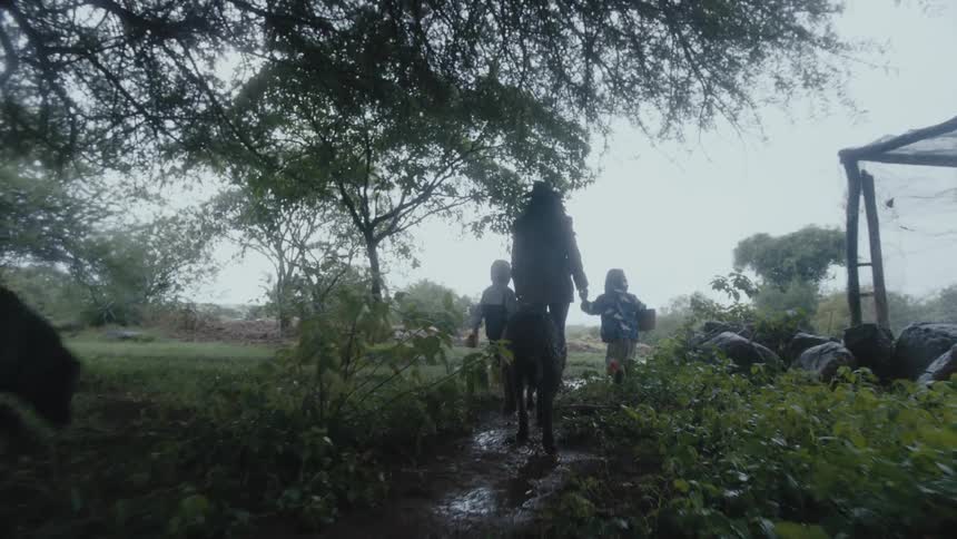 A woman holds her childrens hands as they walk on a paved path near a covered garden