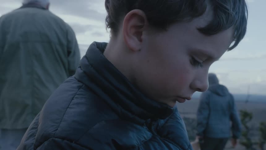 Boy with sad expression looking down and then looking at his grandparents walking at the background