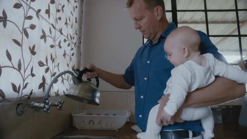 A dad holds his baby on his hip while filling a kettle with water from the kitchen faucet