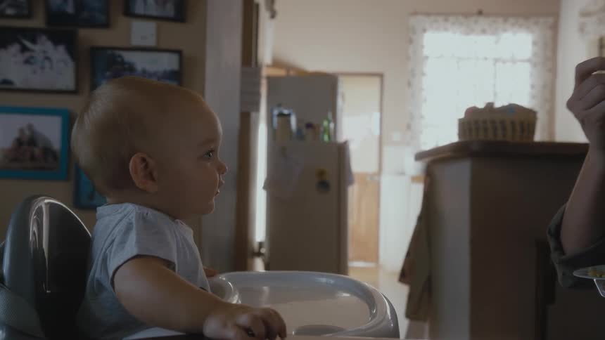Baby sitting at the baby chair next to the kitchen being fed by its mother
