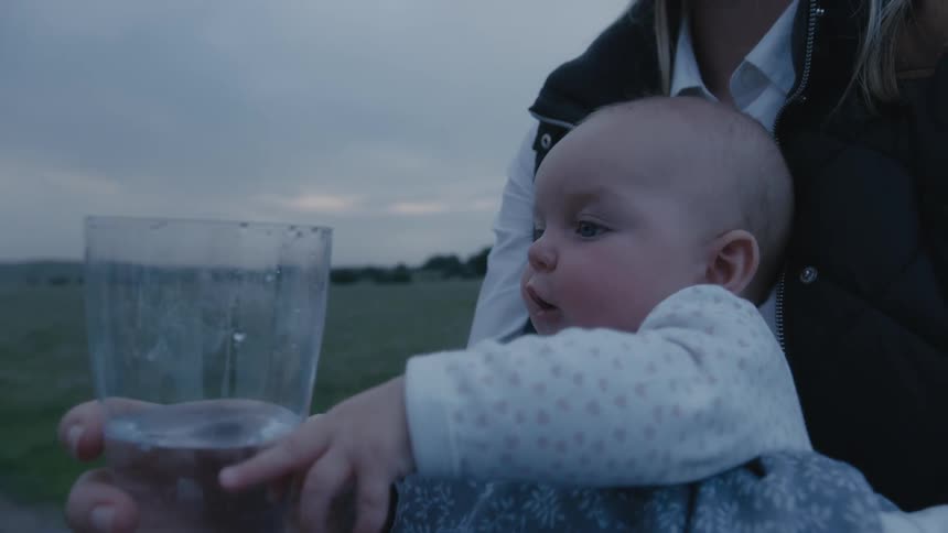 Mother standing in the middle of a grass field holding her baby and a cup of water in front of it