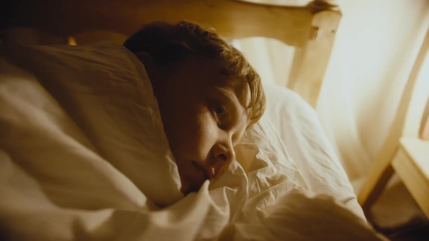 Boy lies in bed looking into space in his room