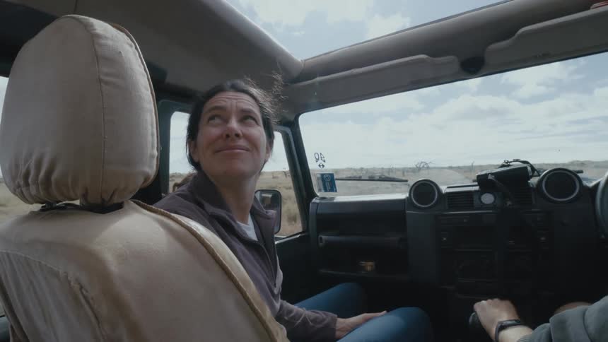 A woman laughs and looks up behind her while riding in an open top jeep through a dirt field