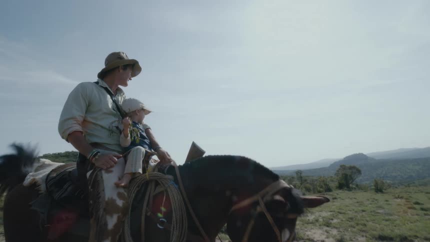 Father and son ride a horse in a grassy field as mountains sit in the distance