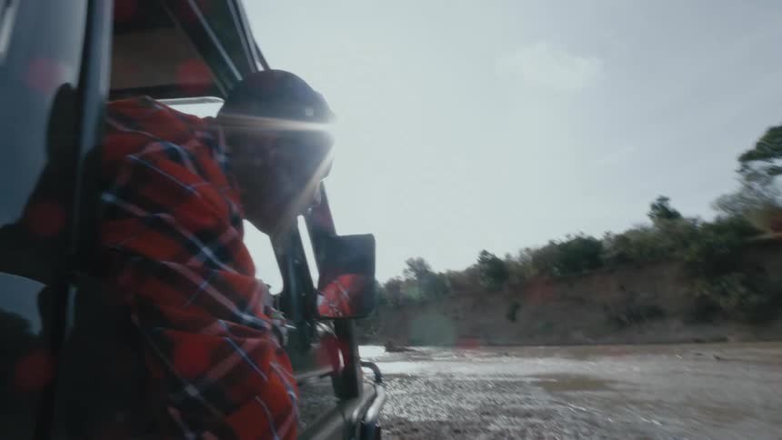 Black man driving a car through a wet terrain near a water stream on a sunny day
