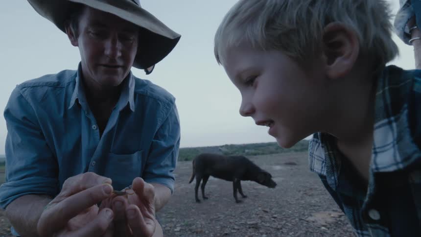 Father kneeling on the ground near a black dog shows his son a small scorpion