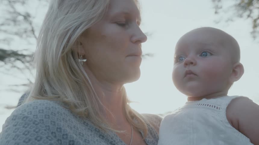 Blonde mother with earrings and shirt holds blue eyed baby under the shining sun
