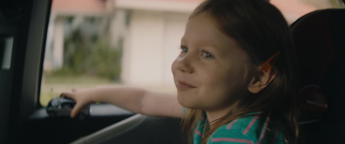 A girl holding a toy car rides in vehicle passing houses in a residential neighborhood