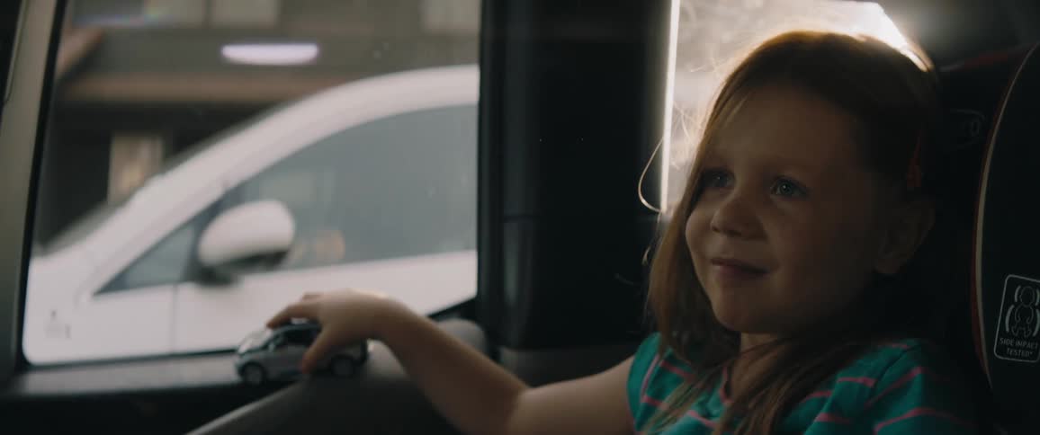 Little girl with turquoise t shirt sitting in car plays with toy car smiling