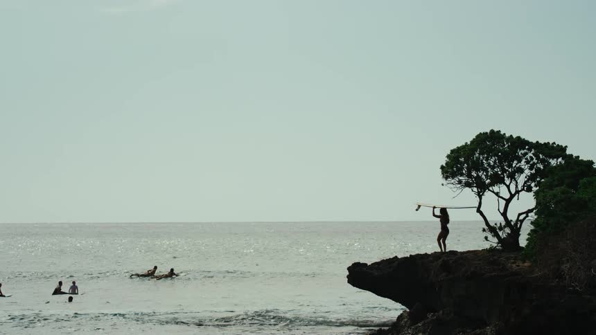 A woman carries a surfboard on her head towards the water where others lie on boards in the water