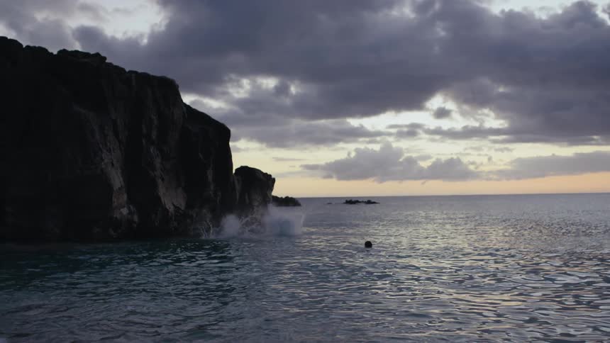 Two people jump off cliff into the sea as red tinted horizon shows beneath a heavy cloudy sky in the background