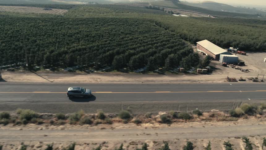 Car travels on the highway and another car is parked on the shoulder near a vineyard as mountains sit in the distance