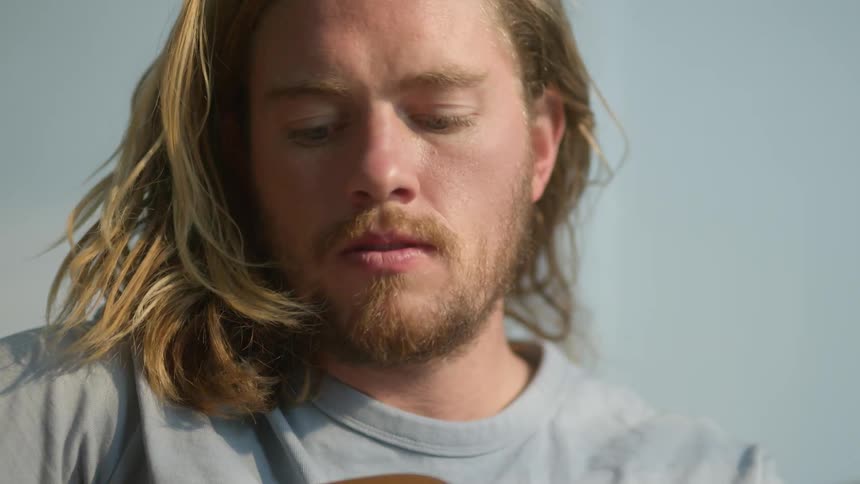 A man with shoulder length blonde hair plays the guitar in the sun