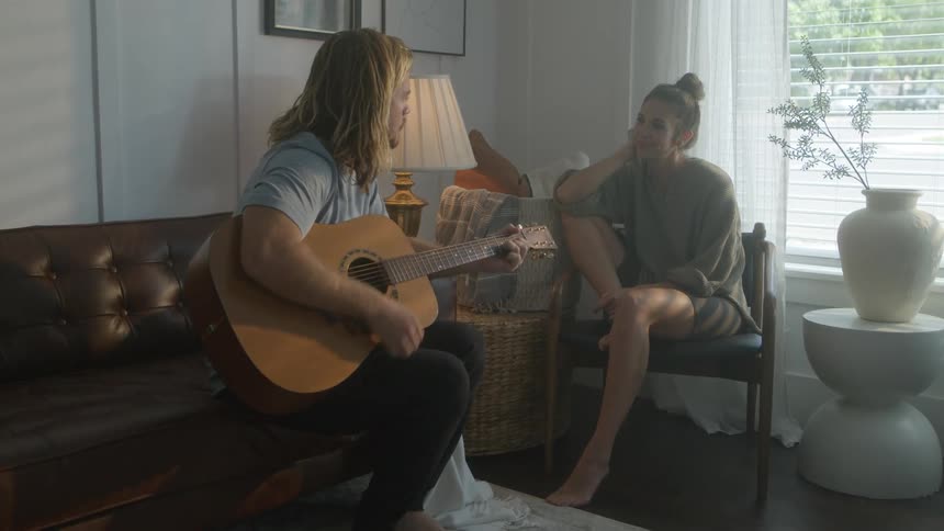 Woman sits with man in living room listening to him playing an acoustic guitar