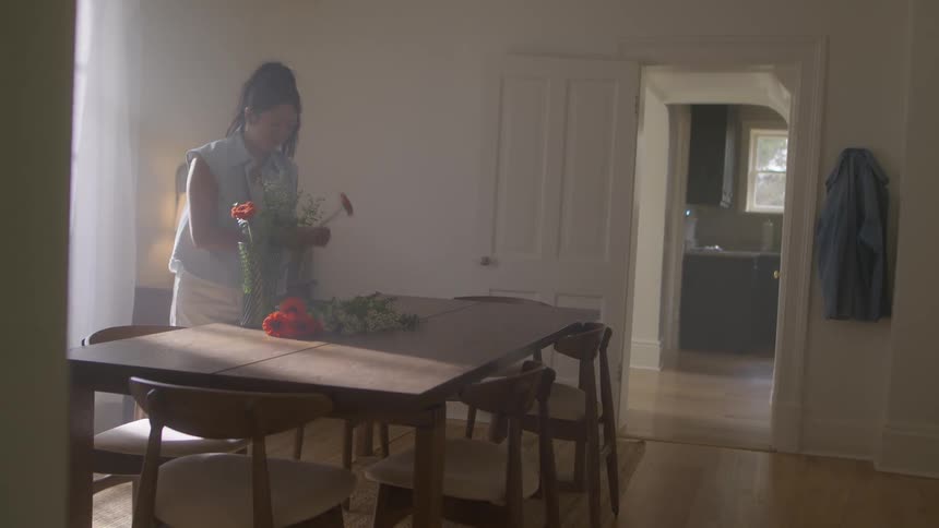 A woman standing in the dining room arranging cut flowers when her husband slides into the room making her laugh