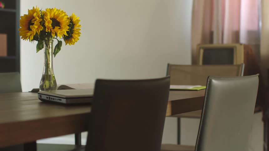 Sunflowers are in a vase on a table with chairs around it and a laptop computer and an open book are also on the table