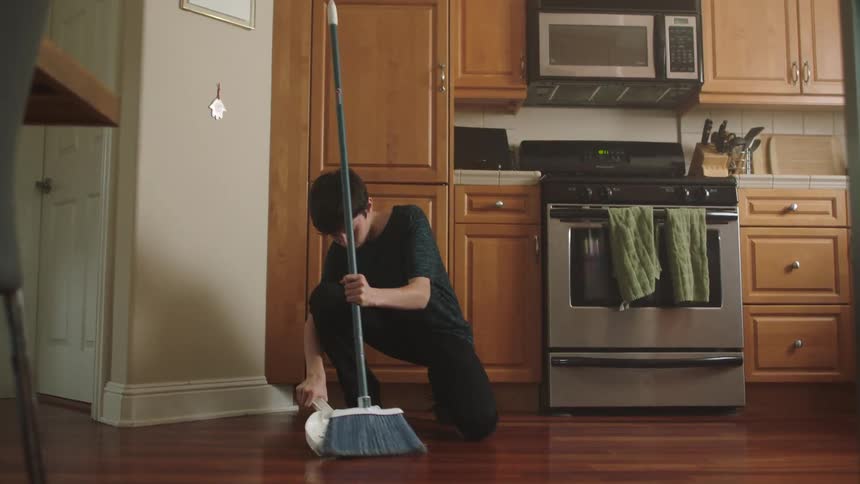 A boy kneeling on the kitchen hardwood floor cleans using a broom and dustpan