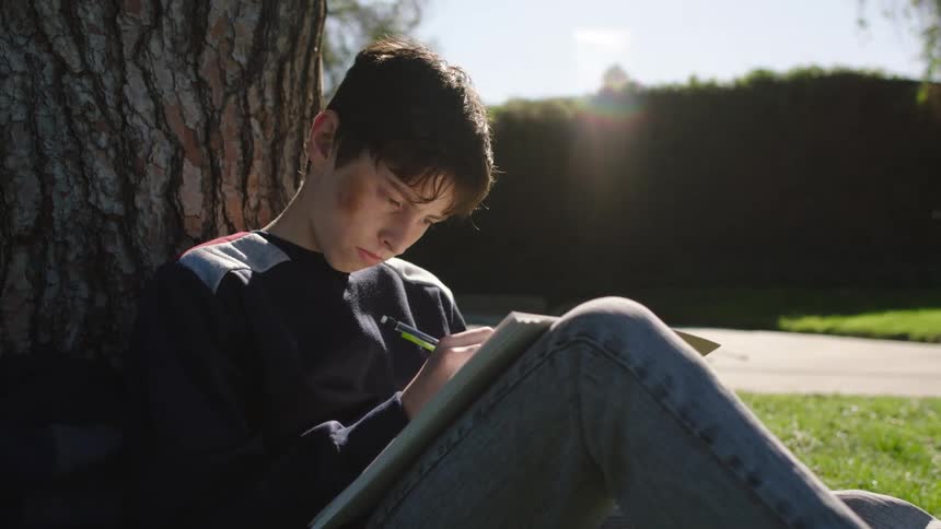A boy sitting against a tree writing in a notebook looks up and leans back