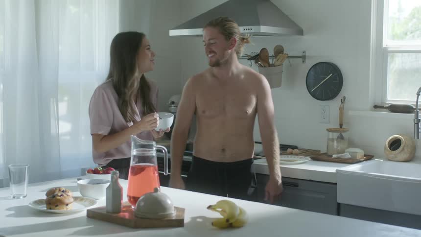 A man cooking at the stove shirtless while his wife eats from a bowl