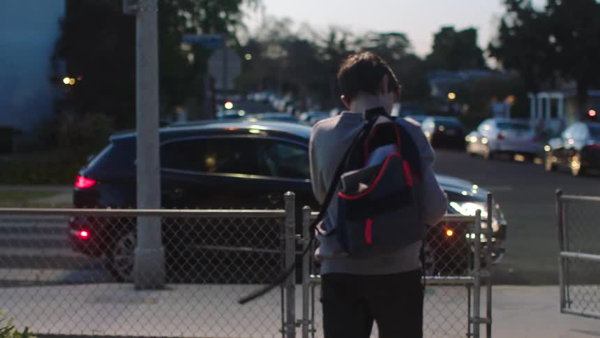 Youth wearing backpack trudges out of gate as car with lit headlights reaches intersection