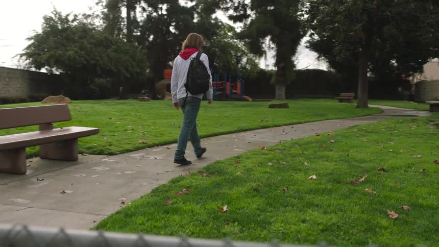 Man carrying backpack enters small park between houses that has a small playground