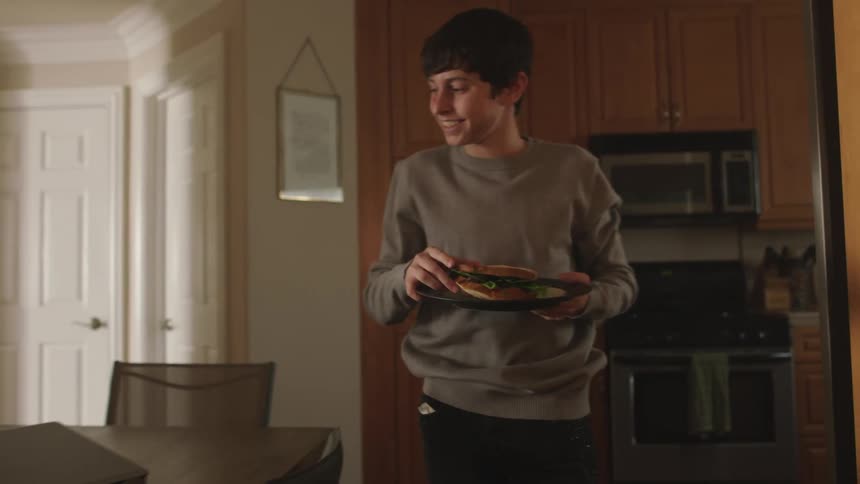Teenaged boy carries a sandwich on a plate to the kitchen table and sits down while looking at a laptop on the table and smiling