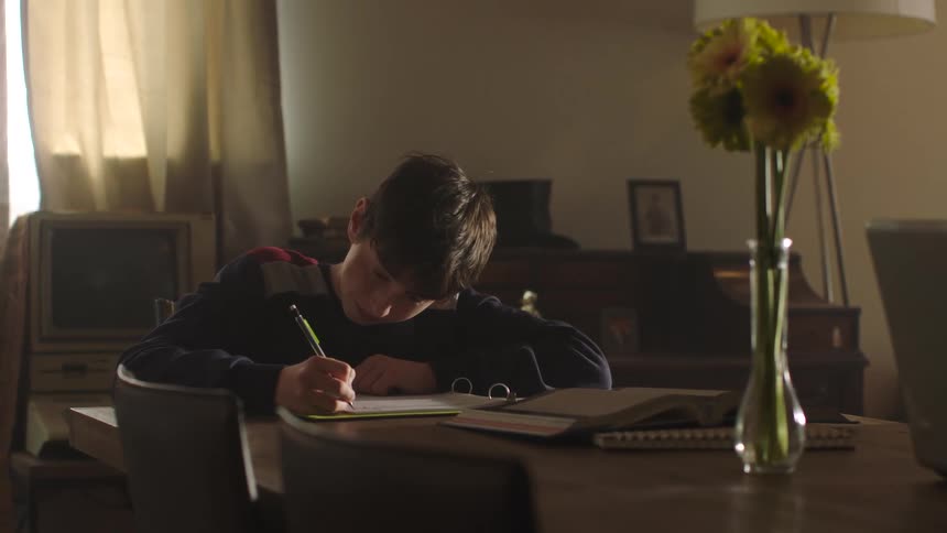A boy is sitting at a table writing in a notebook and he lies his head on the table