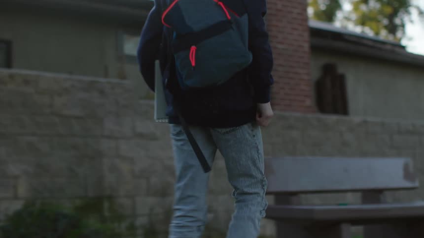 A boy picks up a backpack that is sitting near a tree and walks past houses