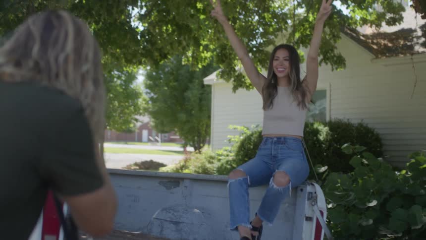A woman is sitting on the back of a white pickup truck posing for a picture as a man with long blond hair holds a camera