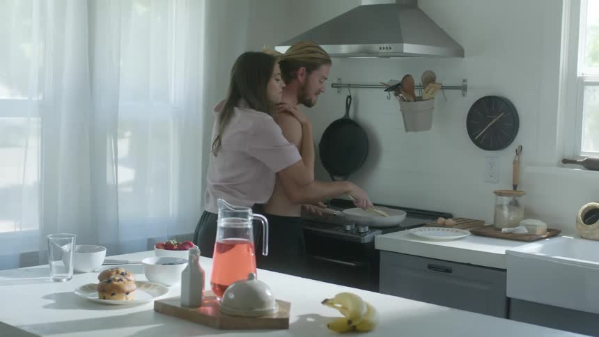 Car passes on street outside nearby window as couple talk while preparing and eating breakfast