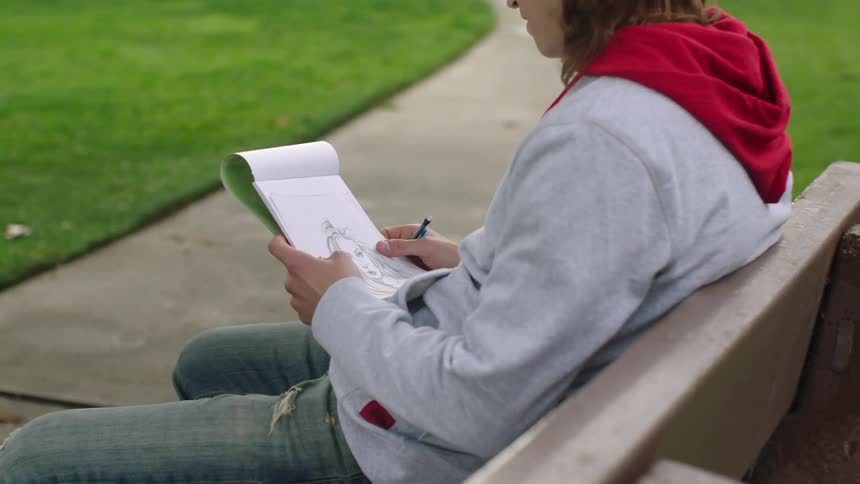 Young man sits on a bench and looks at a sketch of a woman in his notebook