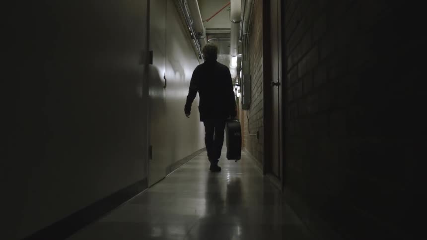 A man is walking down a hall carrying a guitar case