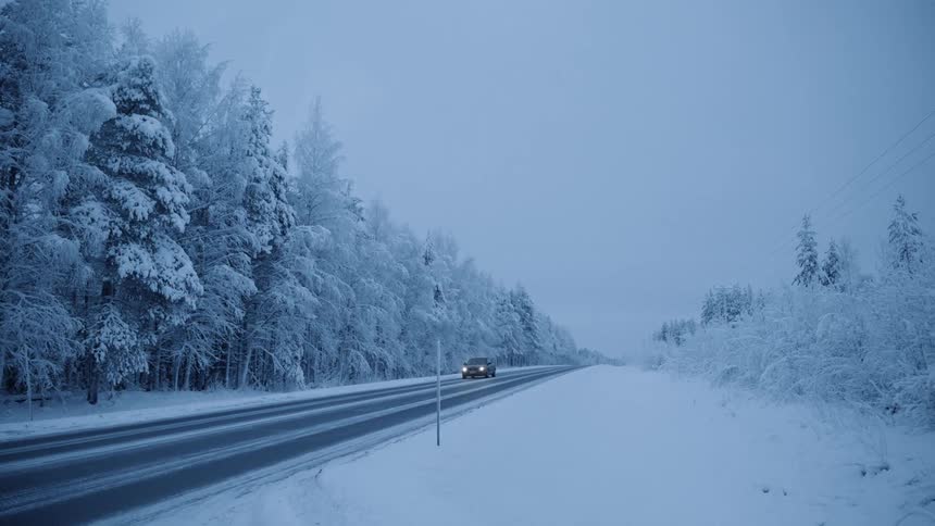 Car travels down the highway in the winter with snow covered trees on one side of the road and snow covered land on the other side