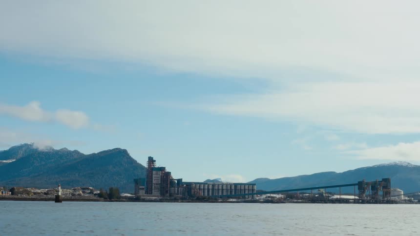 Mountains and a factory and a bridge sit next to the river under the white clouds