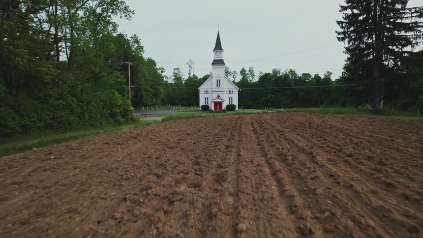 A plowed field leading to a church beside a road in a rural area on a cloudy day