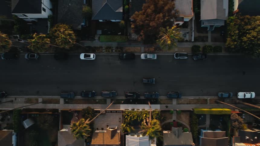 Aerial view of a neighborhood street with cars parked on the roadside in front of houses and palm trees near the sidewalk