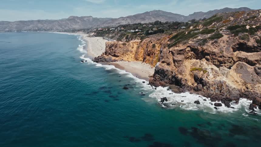 Clear water waves roll over a beach and splash on the rocks near the promontory and coastal community with mountains in the distance