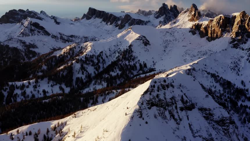 The sun shines brightly upon snow covered mountains with peaks high among the clouds in the sky