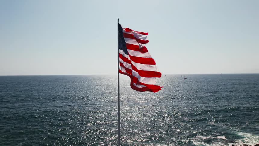 The sun shines on an american flag flying over the water and waving in the wind